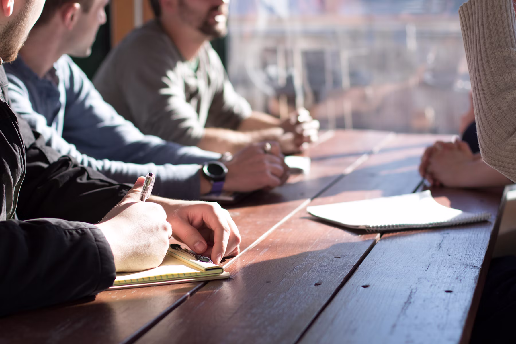 Colleagues with notebooks and pens gathered around a table during a daytime meeting.